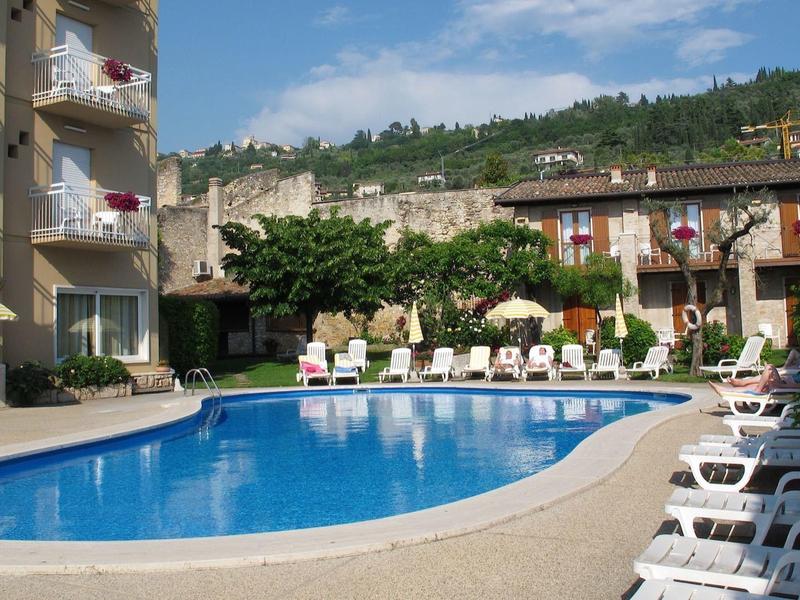 Hotelowy basen otoczony leżakami, budynkami i zielonymi wzgórzami w tle.