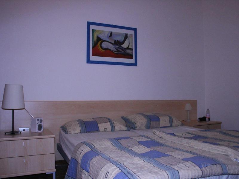 Prosty pokój hotelowy z dwoma łóżkami, stolikiem nocnym i abstrakcyjnym obrazem na ścianie.