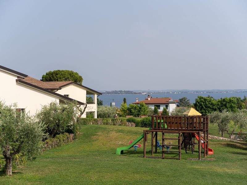Gran jardín con zona de juegos de madera junto a casas blancas y olivos bajo cielo azul.