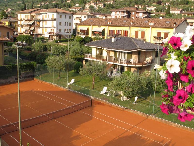 Zona de pista de tenis de tierra roja junto a casas residenciales con flores en primer plano.