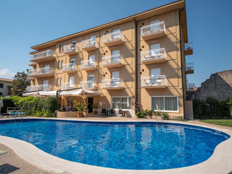 Edificio de hotel de varios pisos con balcones y piscina azul en primer plano bajo cielo despejado.