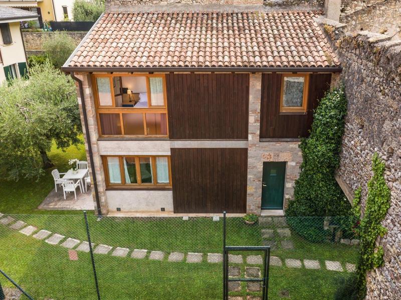 Pequeña casa de dos pisos con techo de tejas y jardín verde junto a un muro de piedra.