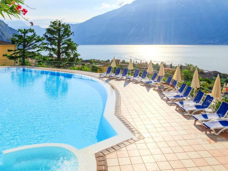 Basen hotelowy z leżakami z widokiem na jezioro i góry w słoneczny dzień.