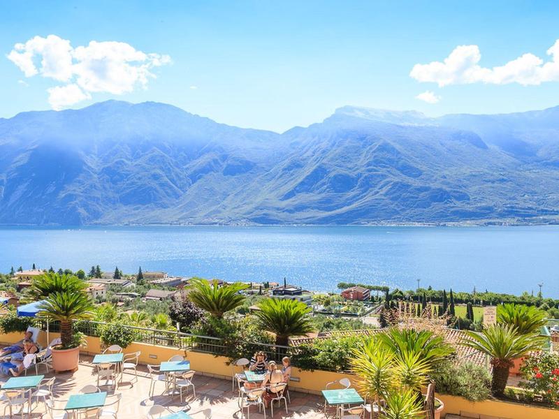 Widok na taras z stolikami i roślinami z widokiem na jezioro i góry w tle.