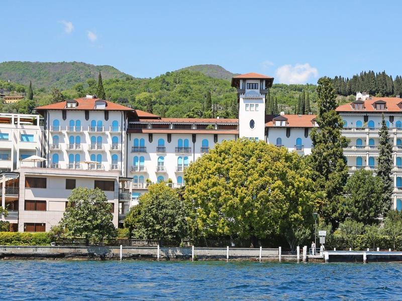 Grand hôtel avec de nombreuses fenêtres devant une montagne boisée et un ciel bleu.