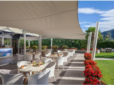 Area esterna di un hotel con tavoli apparecchiati, sedie bianche, fiori rossi e tende da sole.