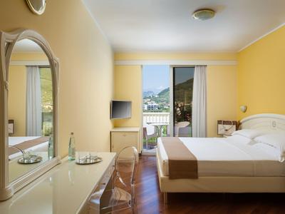 Camera d'hotel luminosa con letto matrimoniale, pareti gialle, TV a schermo piatto e balcone con vista sulle montagne.