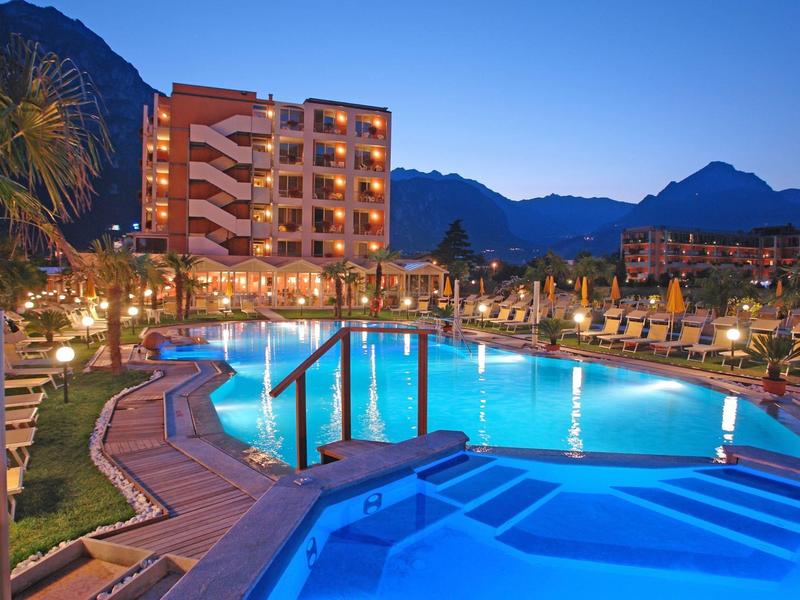 Evening view of a lit hotel pool with loungers and mountains in the background.