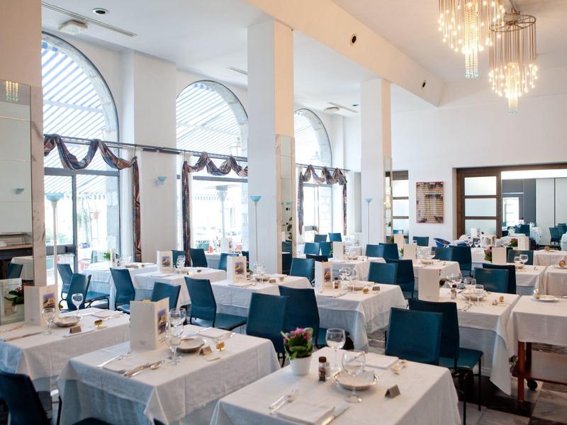 Licht, elegant restaurant met witte tafelkleden, blauwe stoelen en kroonluchters.