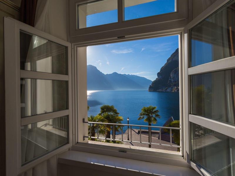 Uitzicht vanuit een open raam op een meer en bergen in zonlicht.