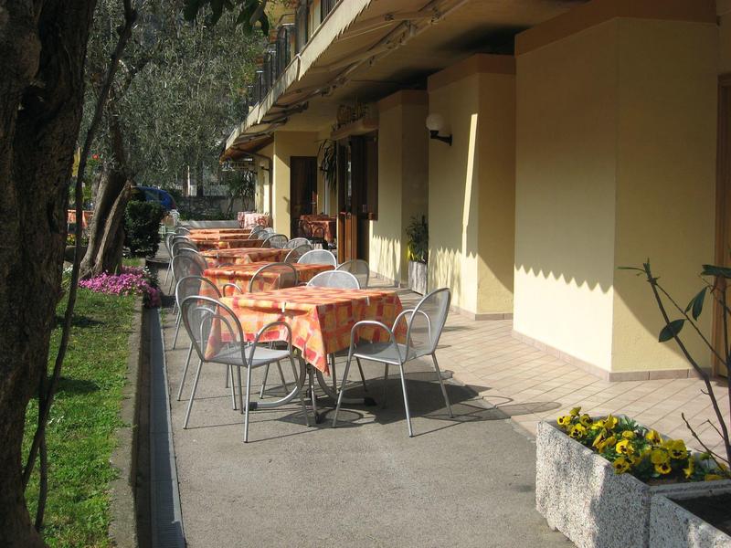 Terrasse mit Tischen und Stühlen, weiße Metallstühle, orange karierte Tischtücher, Blumendekor.