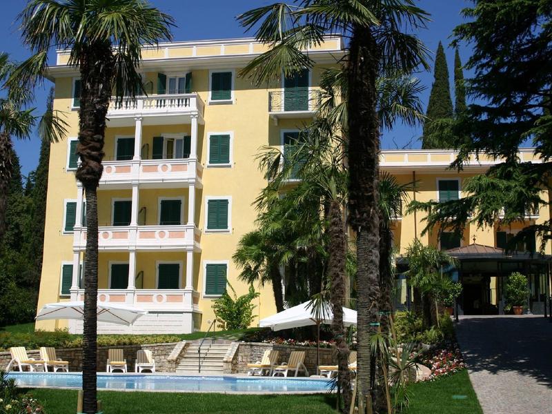 Geel hotel met vier verdiepingen en balkons, omgeven door palmbomen en ligstoelen bij het zwembad.