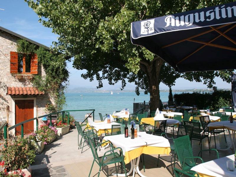 Terrassenrestaurant mit gelben Tischdecken, grünen Stühlen, Bäumen und Blick auf einen See.