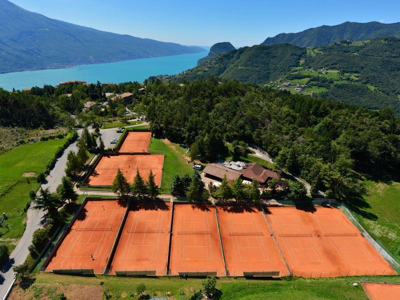 Vue sur des courts de tennis en terre battue rouge dans une région montagneuse verte près d'un lac.