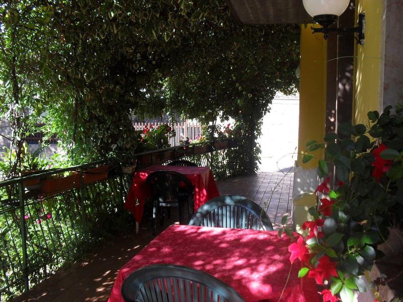 Area esterna con tavoli rossi e sedie verdi accanto a parete gialla e fiori.