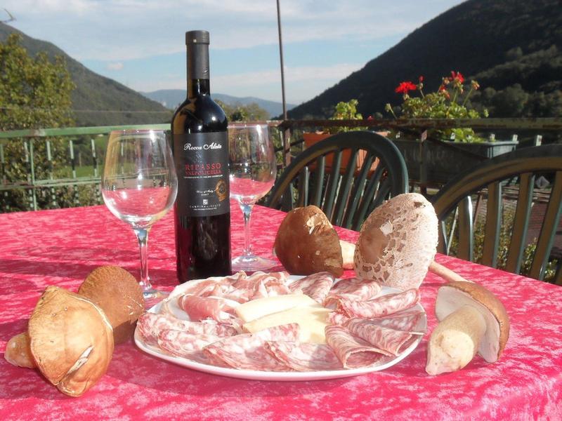 Terrazza con tavolo apparecchiato con vino, bicchieri, pane e salumi davanti a uno sfondo montano.