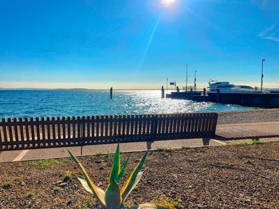 Pohled na pobřeží s lavičkou v popředí a lodí u mola za jasné oblohy.
