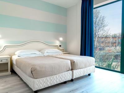 Světle hotelový pokoj se dvěma jednolůžky, nočním stolkem a velkým panoramatickým oknem s závěsem.