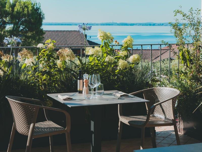 Terasa se stolem a čtyřmi židlemi před kvetoucími rostlinami a výhledem na jezero.