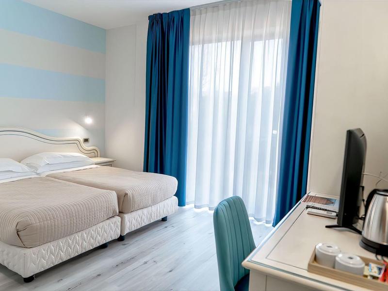 Světle hotelový pokoj se dvěma jednolůžky, psacím stolem a závěsy u okna.