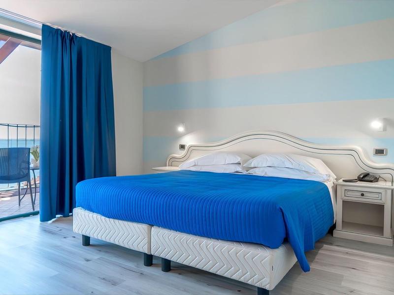 Moderní hotelový pokoj s modrou postelí a přístupem na balkon s posezením.