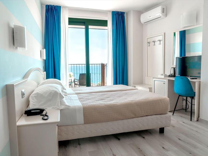 Moderní hotelový pokoj s manželskou postelí, modrobílou výzdobou a balkonem s výhledem na moře.