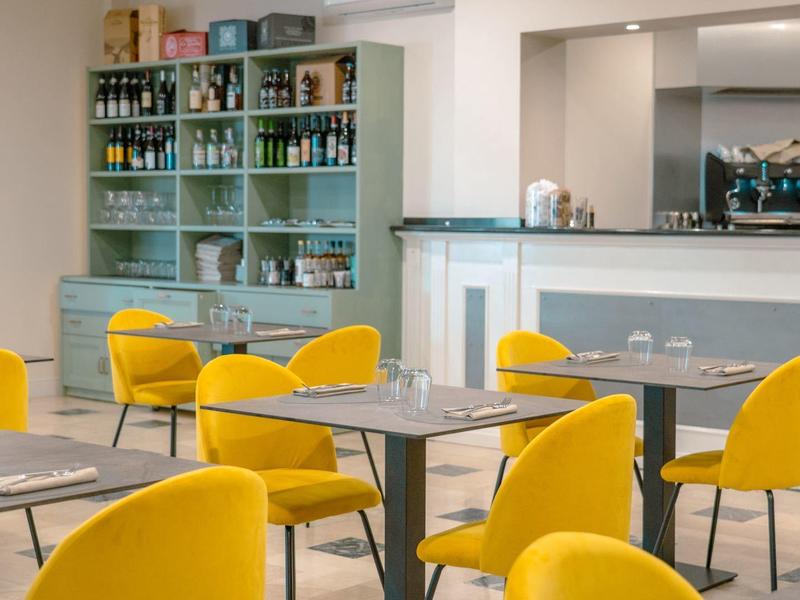 Moderní kavárna se žlutými židlemi, šedými stoly a regály s nápoji v pozadí.