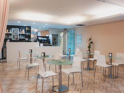 Jasna kawiarnia z okrągłymi stołami, białymi krzesłami i barem z napojami w tle.