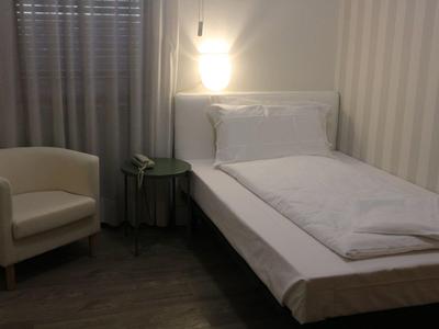 Una semplice camera singola con letto bianco, comodino e poltrona beige accanto a una finestra con tende.