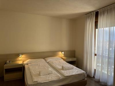 Eenvoudige hotelkamer met een tweepersoonsbed, nachtkastjes en lichte gordijnen bij het raam.