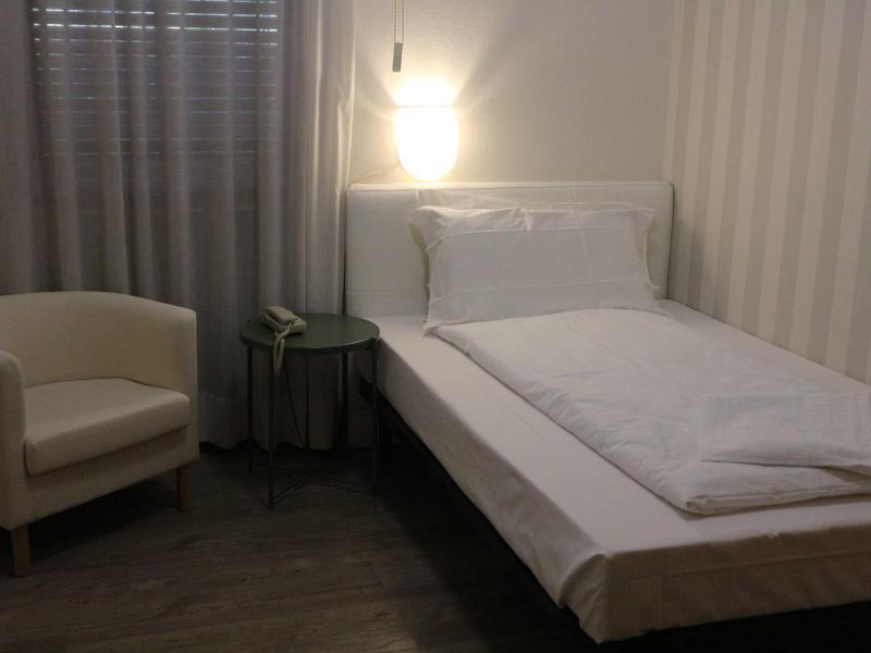 Una semplice camera singola con letto bianco, comodino e poltrona beige accanto a una finestra con tende.
