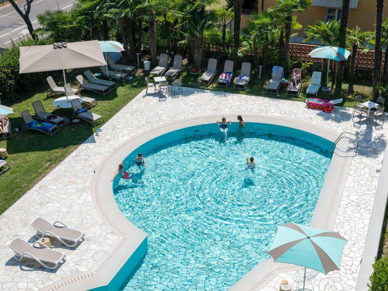 Piscina con ombrelloni e lettini nel giardino verde di un hotel.