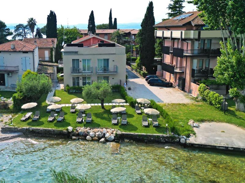 Zelený travnatý břeh jezera s lehátky a slunečníky před domy.