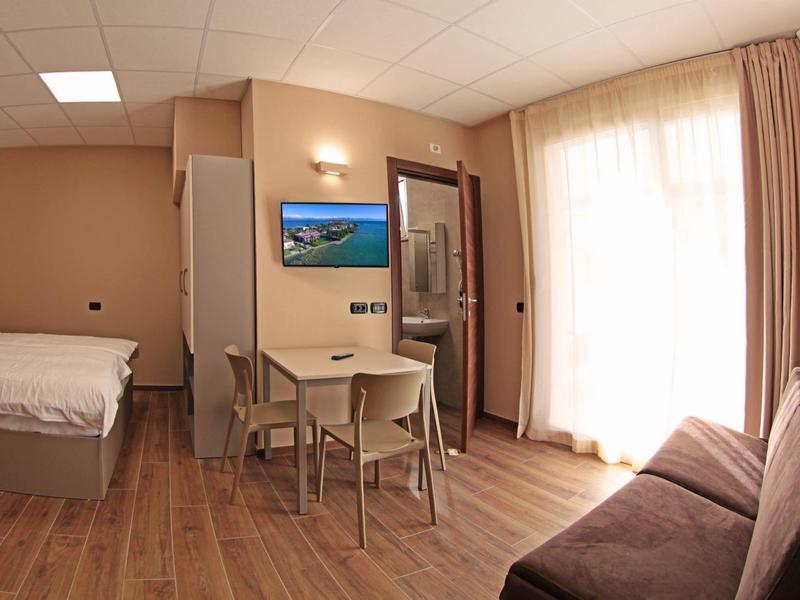 Moderně zařízený hotelový pokoj s manželskou postelí, jídelním stolem, pohovkou a přístupem na balkon.