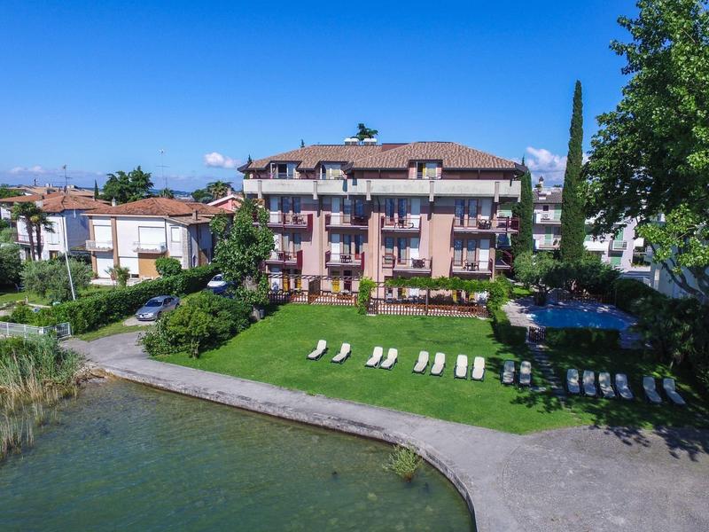 Hotel with garden area, sun loungers, and direct access to a lake under a blue sky.