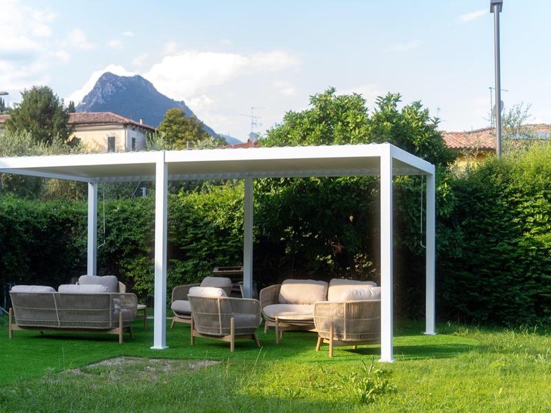 Padiglione moderno con mobili da giardino su prato verde davanti a siepe e montagne sullo sfondo.