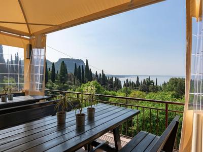 Terrazza con tavolo in legno e panchine sotto tenda con vista su paesaggio verde e mare.