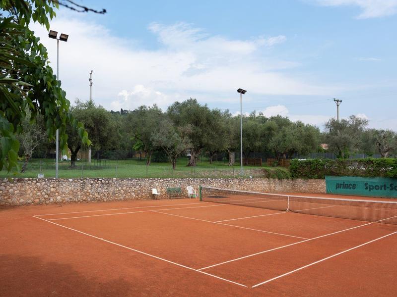 Campo da tennis in terra rossa con alberi verdi e cielo grigio-azzurro sullo sfondo.
