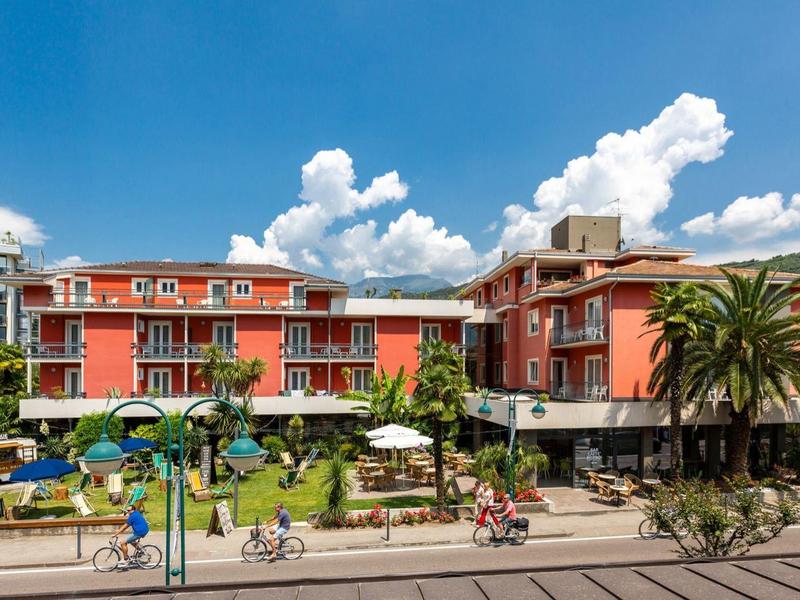 Rotes Hotelgebäude mit Balkon, Palmen, Pool und Sonnenschirmen unter blauem Himmel.