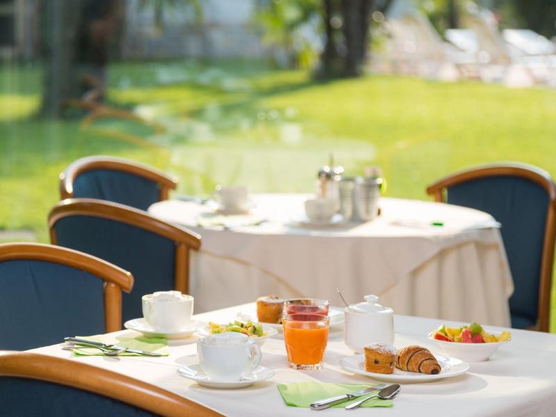 Mehrere gedeckte Tische mit weißen Tischdecken, Frühstücksgeschirr, Croissants und Orangensaft im Garten.