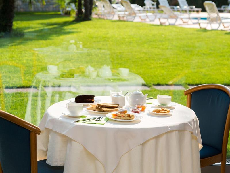 Runder Tisch mit weißer Tischdecke und dampfenden Tellern steht vor grüner Wiese mit Liegestühlen.