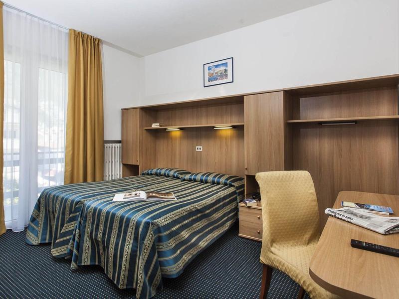 Ein einfaches Hotelzimmer mit zwei Betten, Holzmöbeln, gelben Vorhängen und Teppichboden.