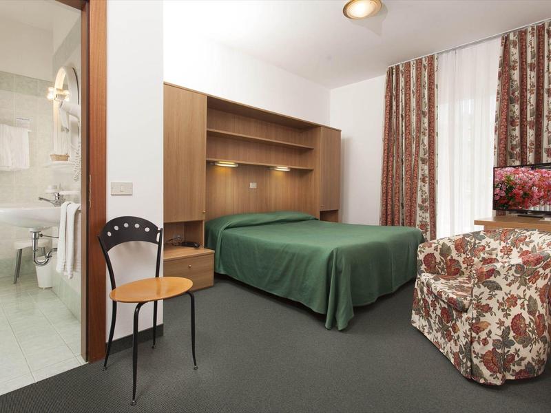 Einfaches Hotelzimmer mit grünem Bett, Holzregal, floralem Sessel und angrenzendem Badezimmer.