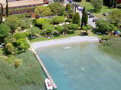 Kleiner Steg am Ufer mit Boot, grüne Bäume, Gras und Gebäude im Hintergrund.