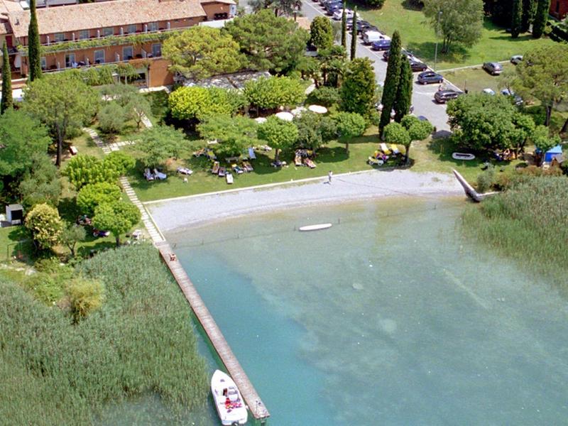Kleiner Steg am Ufer mit Boot, grüne Bäume, Gras und Gebäude im Hintergrund.