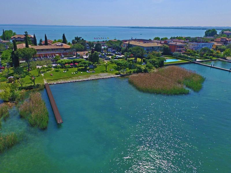 Klares Wasser, grüne Ufer mit Pflanzen, Gebäude und blauer Himmel über ruhigem See.