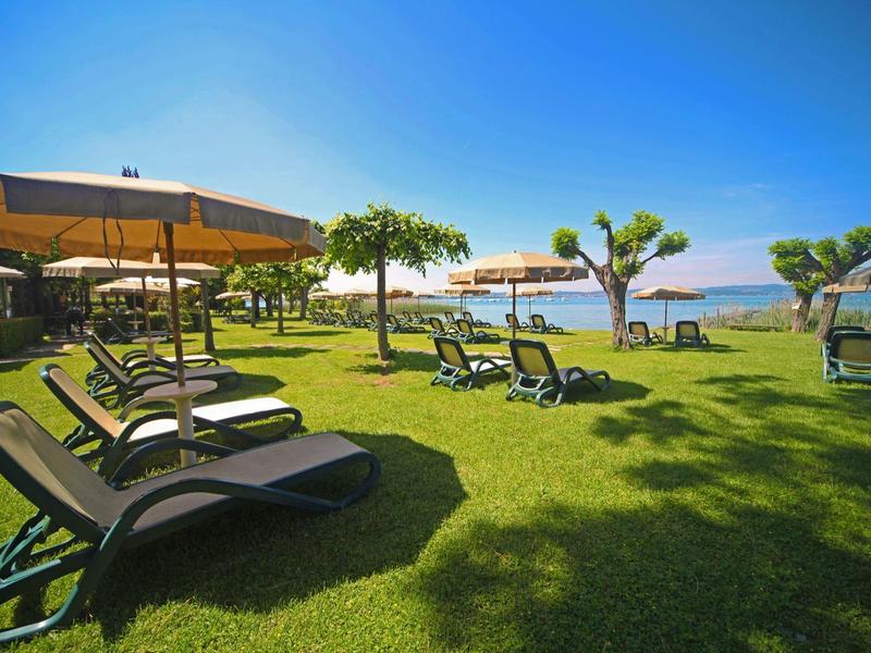 Grüne Wiese mit Liegestühlen und Sonnenschirmen am Meer unter klarem blauem Himmel.