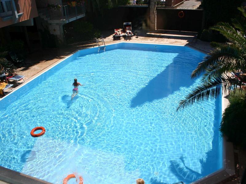 Großer Außenpool mit klarem blauem Wasser, umgeben von Liegestühlen und Palmen, Person schwimmt.
