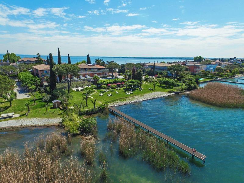 Grüne Uferlandschaft mit Bäumen, Häusern und einem Steg am ruhigen, blauen See unter blauem Himmel.