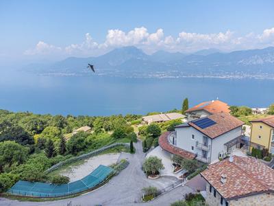 Vista di case e colline boscose con lago e montagne sullo sfondo sotto un cielo limpido.